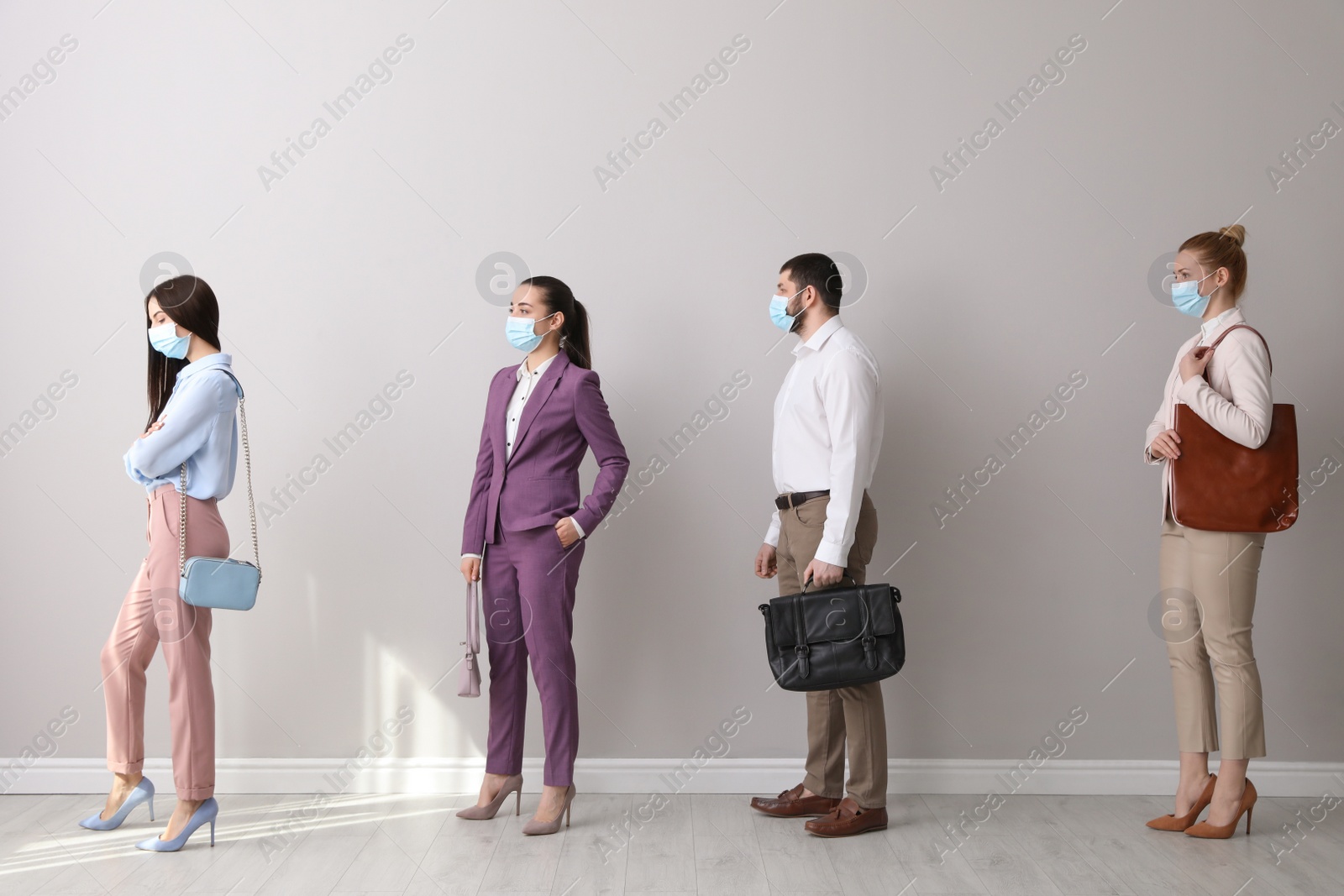 Photo of Businesspeople with protective masks waiting in queue near light wall indoors. Social distancing during Covid-19 pandemic