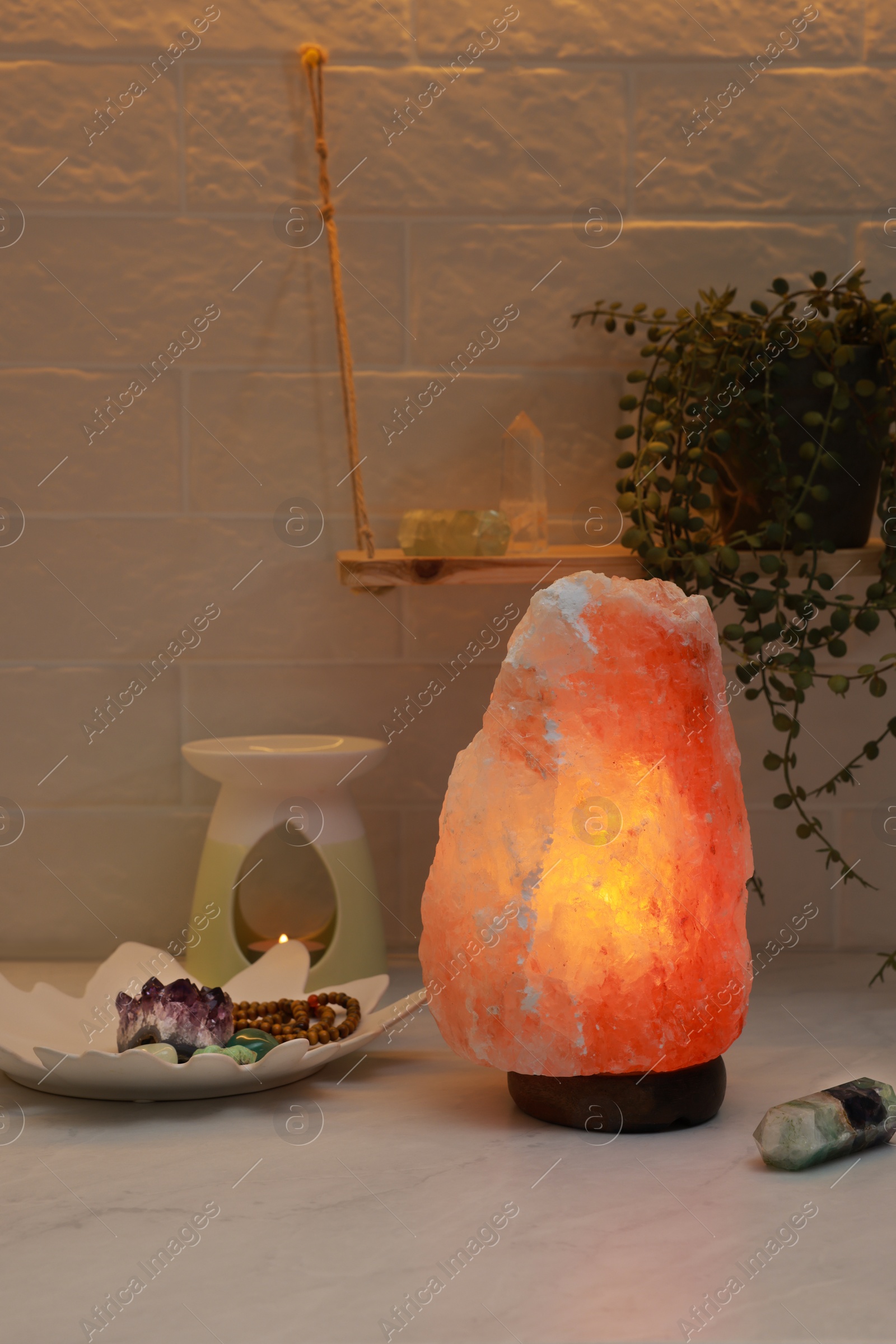 Photo of Himalayan salt lamp, crystals and oil diffuser on white table near brick wall