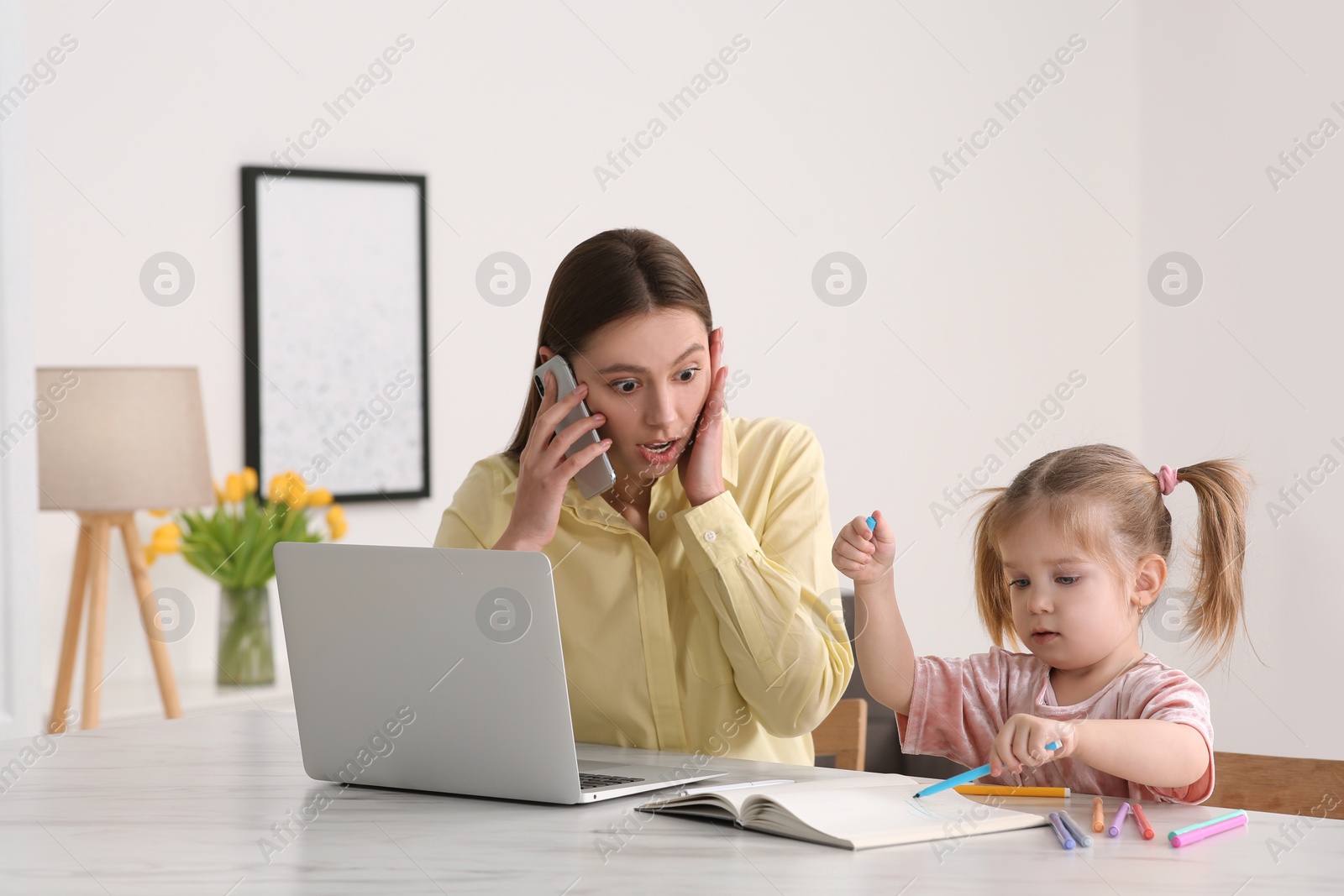 Photo of Woman working remotely at home. Shocked mother looking while daughter drawing in notebook at desk