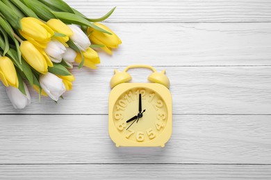 Yellow alarm clock and beautiful tulips on white wooden table, flat lay. Spring time