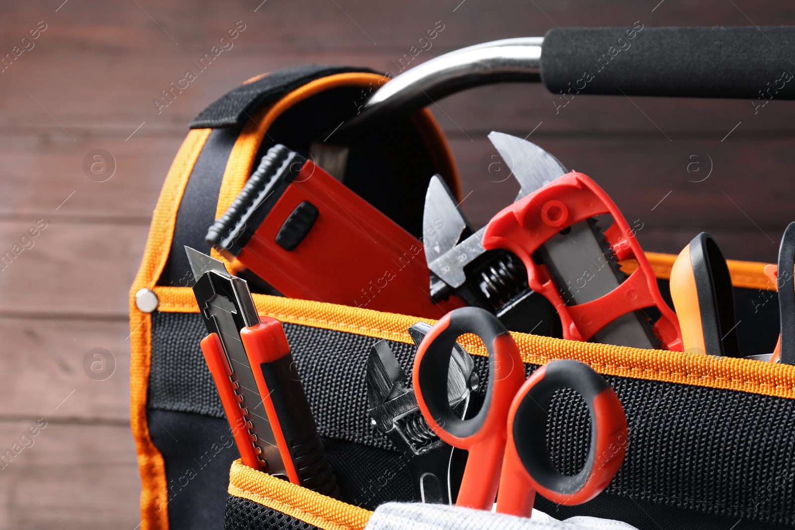 Photo of Bag with utility knife and different tools on wooden background, closeup