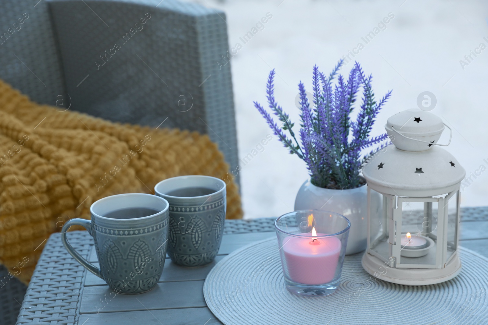 Photo of Burning candle, lantern, potted flowers and cups with hot drink on coffee table outdoors. Cosy winter