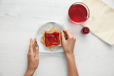 Photo of Woman eating toast with jam at white wooden table, top view. Delicious morning meal