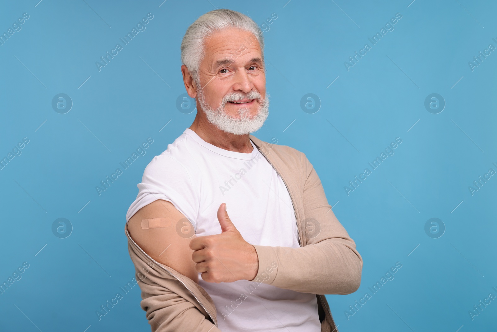 Photo of Senior man with adhesive bandage on his arm after vaccination showing thumb up against light blue background