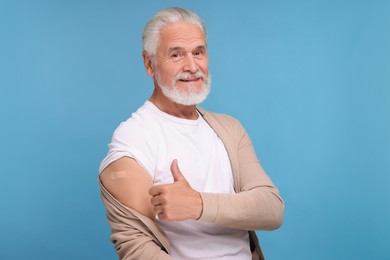 Senior man with adhesive bandage on his arm after vaccination showing thumb up against light blue background