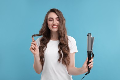 Photo of Happy young woman with beautiful hair holding curling iron on light blue background