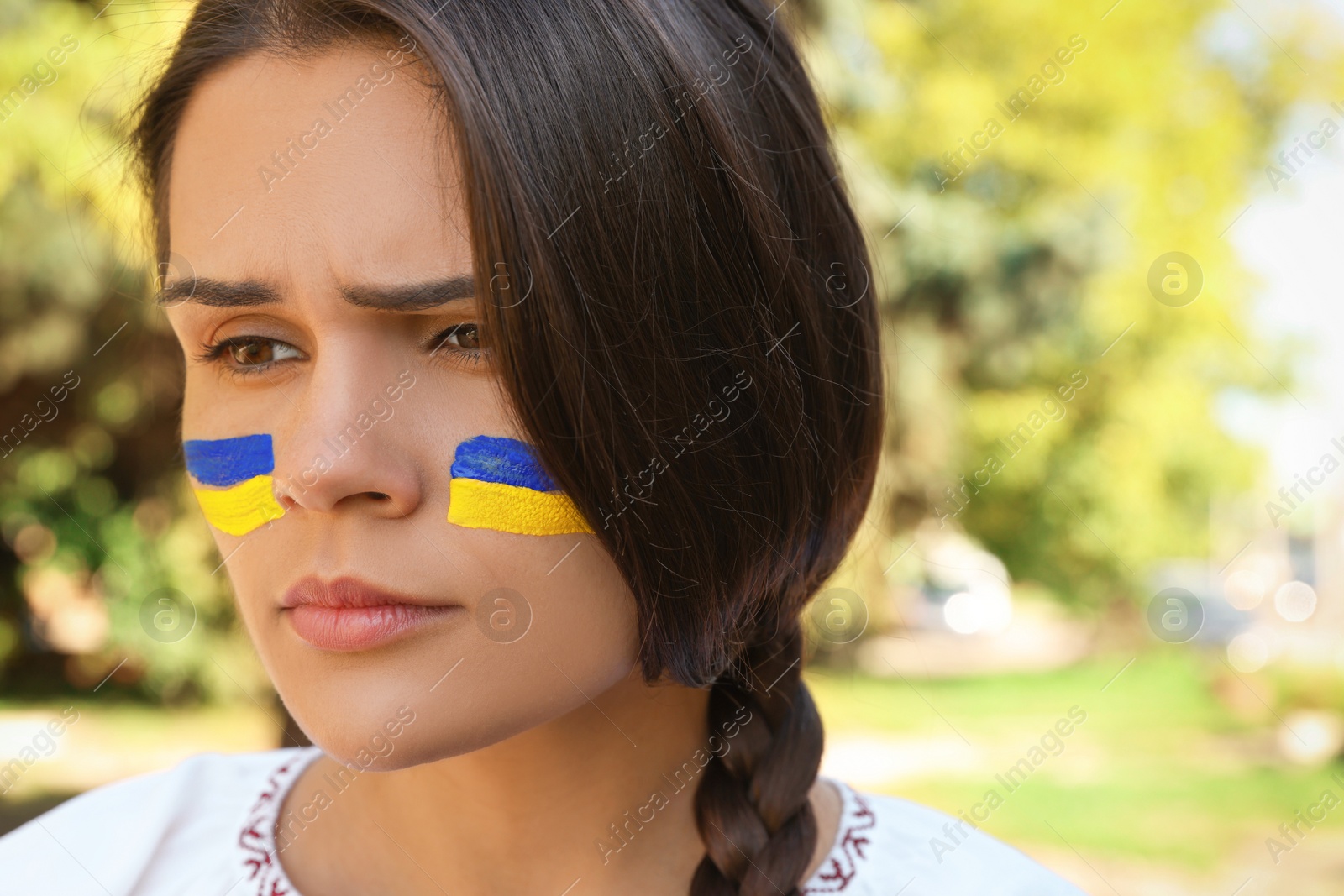 Photo of Sad young woman with drawings of Ukrainian flag on face outdoors, closeup. Space for text