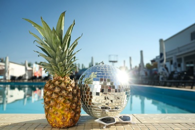 Photo of Shiny disco ball, pineapple and sunglasses on edge of swimming pool. Party items