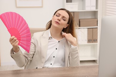 Photo of Businesswoman waving pink hand fan to cool herself at table in office