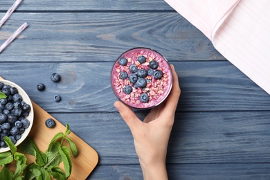 Woman holding glass of delicious blueberry smoothie with granola at blue wooden table, top view