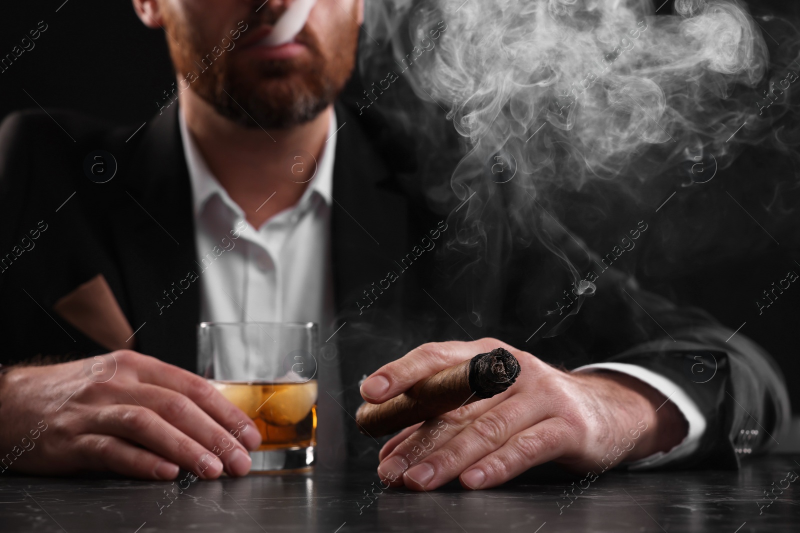 Photo of Man with glass of whiskey smoking cigar at dark marble table on black background, closeup