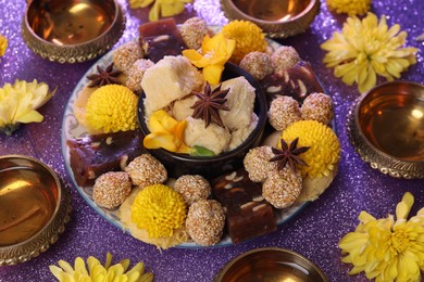 Photo of Diwali celebration. Diya lamps, tasty Indian sweets and yellow flowers on shiny violet table