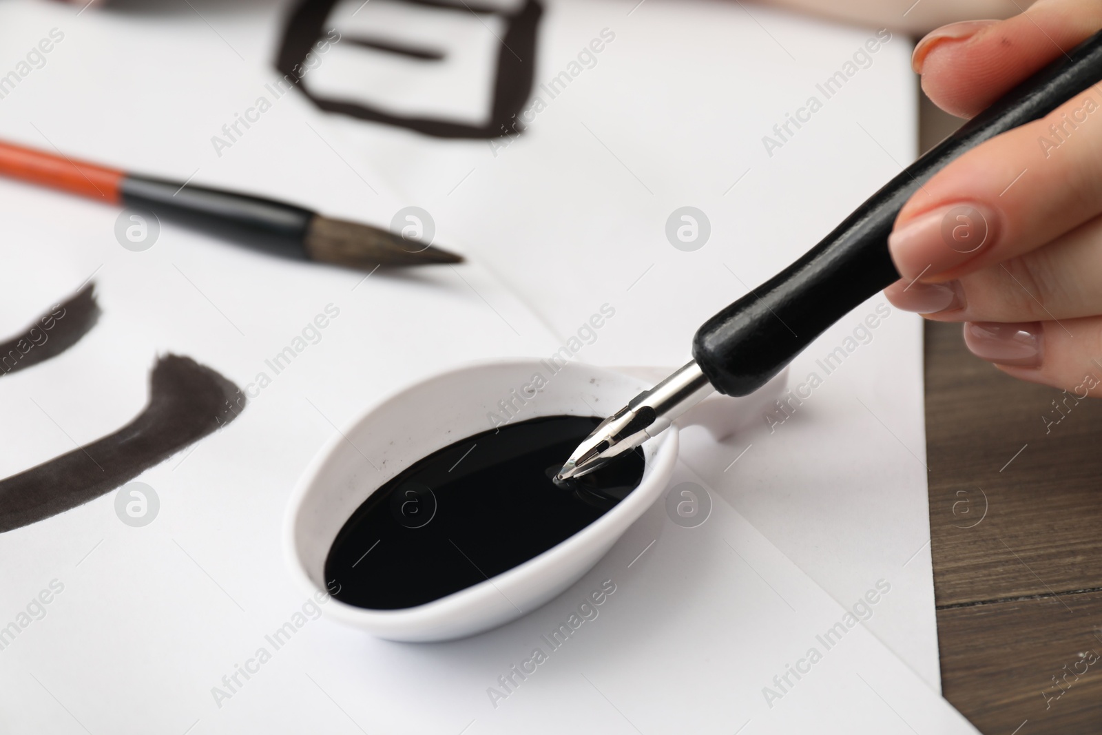 Photo of Calligraphy. Woman dipping brush into inkwell at table, closeup