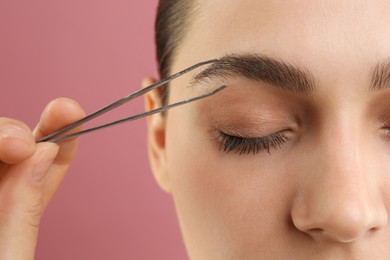 Photo of Eyebrow correction. Young woman with tweezers on pink background, closeup