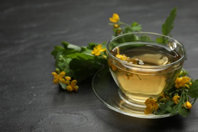 Photo of Glass cup of aromatic celandine tea and flowers on black table. Space for text