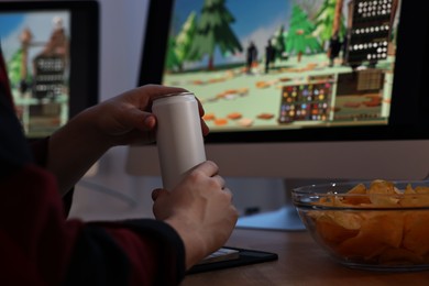 Young man with energy drink and chips playing video game at wooden desk indoors, closeup