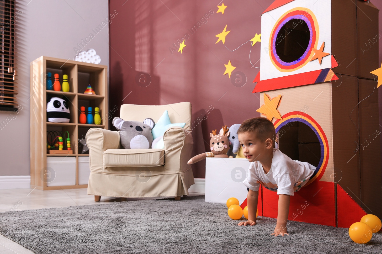 Photo of Cute little boy playing with cardboard rocket and toys at home. Child's room interior