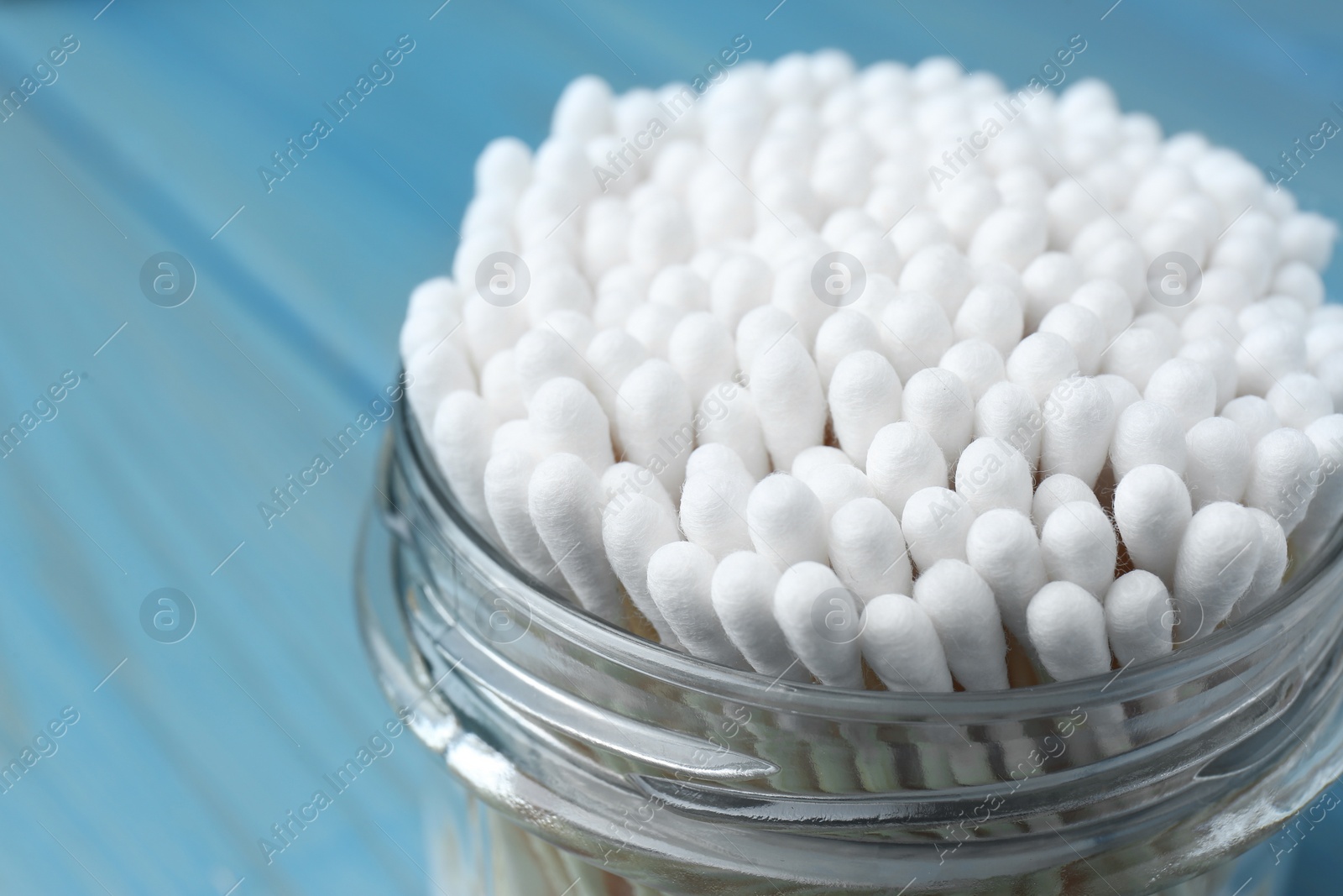 Photo of Many cotton buds in glass jar on light blue background, closeup