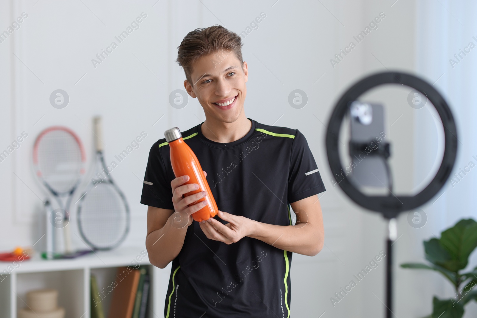 Photo of Smiling sports blogger holding bottle while streaming online fitness lesson with smartphone at home