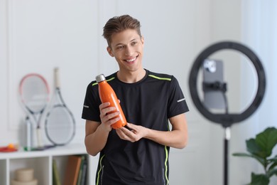 Smiling sports blogger holding bottle while streaming online fitness lesson with smartphone at home