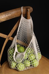 Photo of Green apples in net bag hanging on wooden chair against black background, closeup