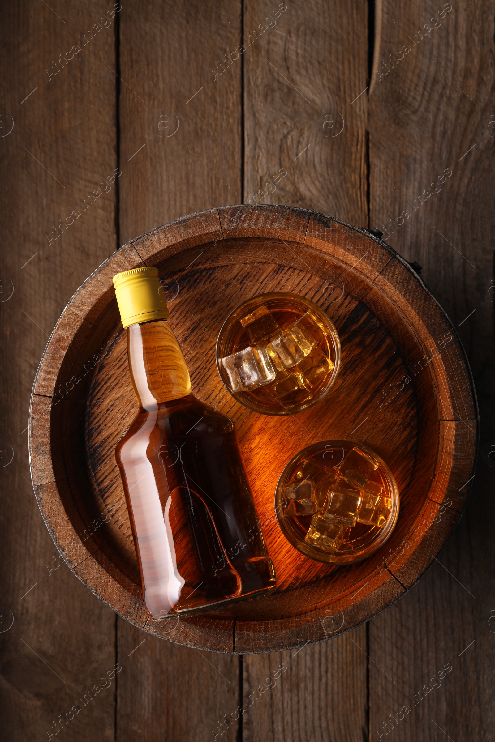 Photo of Whiskey with ice cubes in glasses, bottle and barrel on wooden table, top view
