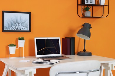 Modern laptop, books, lamp and stationery on wooden desk near orange wall. Home office