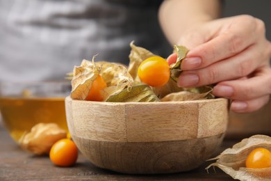 Photo of Woman peeling physalis fruit from calyxes at wooden table, closeup