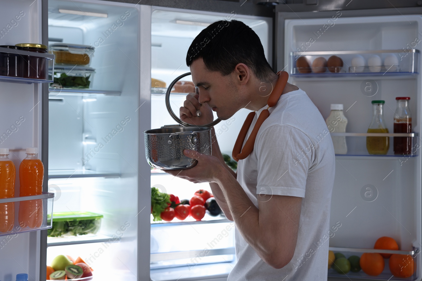 Photo of Man with sausages and pot near refrigerator in kitchen at night