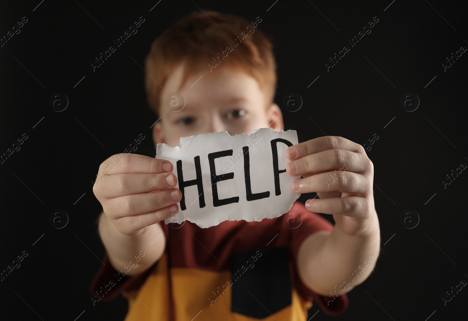 Photo of Little boy holding piece of paper with word Help against black background, focus on hands. Domestic violence concept