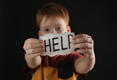 Little boy holding piece of paper with word Help against black background, focus on hands. Domestic violence concept