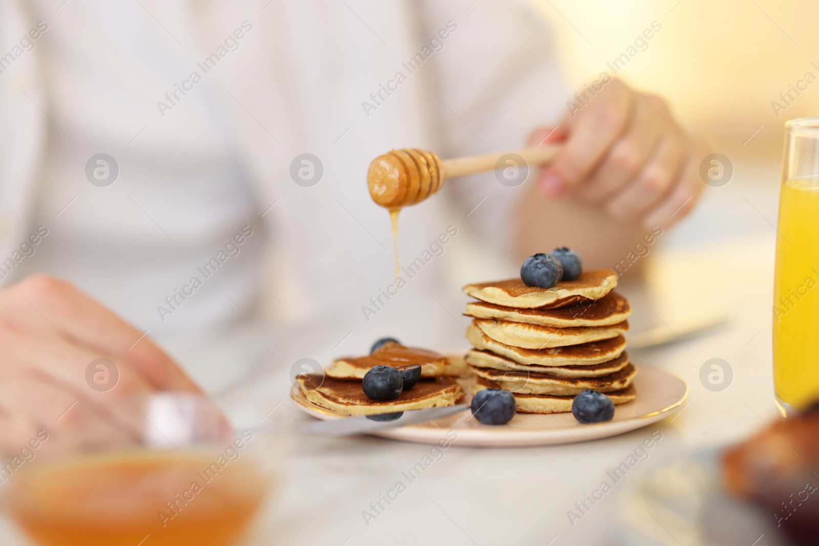 Photo of Man pouring honey onto pancakes with blueberries at table, closeup