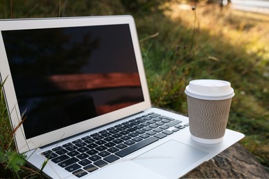 Photo of Modern laptop with blank screen and coffee cup on stone in nature, space for text. Working outdoors
