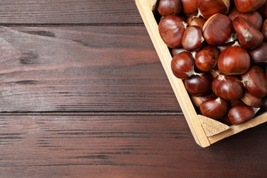 Photo of Fresh sweet edible chestnuts in crate on wooden table, top view. Space for text