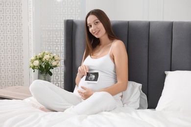 Photo of Pregnant woman with ultrasound picture of baby on bed indoors