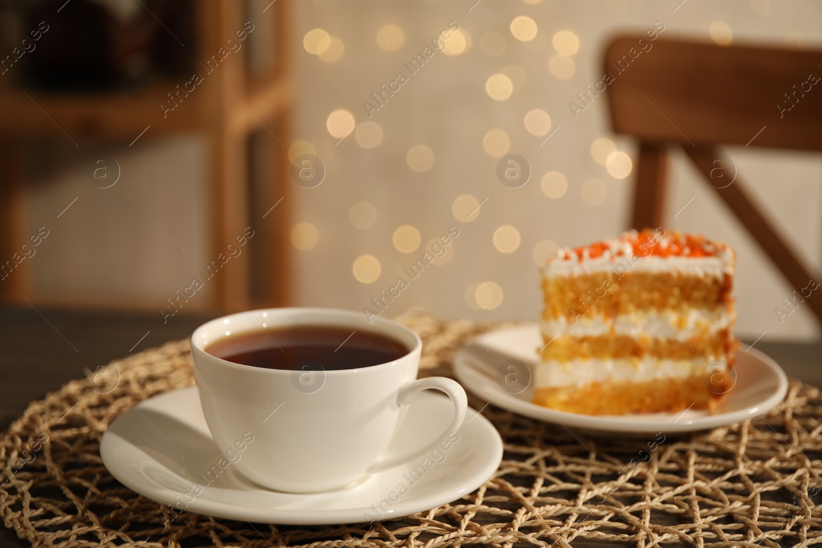 Photo of Hot tea in cup and piece of cake on wooden table against blurred lights indoors