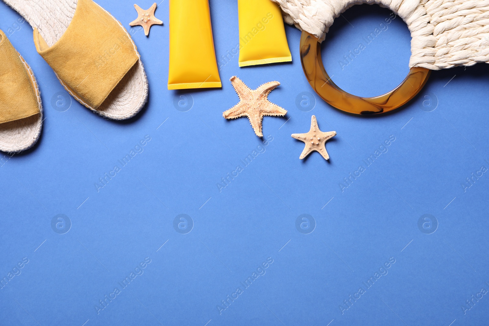 Photo of Flat lay composition with slippers, bag and sun protection cosmetic products on blue background. Space for text