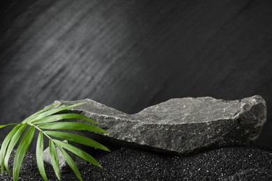 Photo of Presentation of product. Stone podium and palm leaves on black sand against dark textured background. Space for text