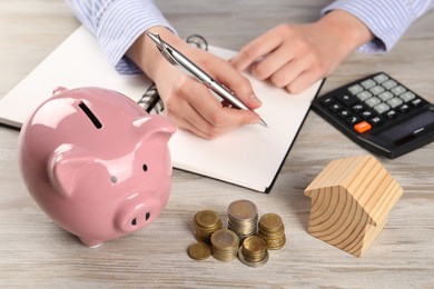 Woman planning budget at wooden table, focus on house model, coins and piggy bank