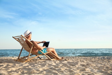 Young man with laptop in deck chair on beach