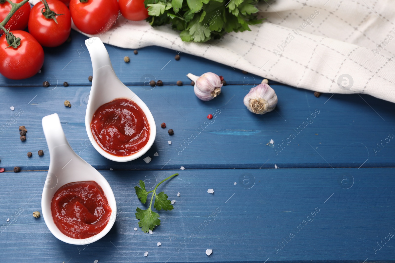 Photo of Organic ketchup in spoons, fresh tomatoes and spices on blue wooden table. Tomato sauce