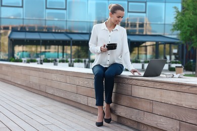 Photo of Smiling businesswoman holding lunch box and working with laptop outdoors. Space for text