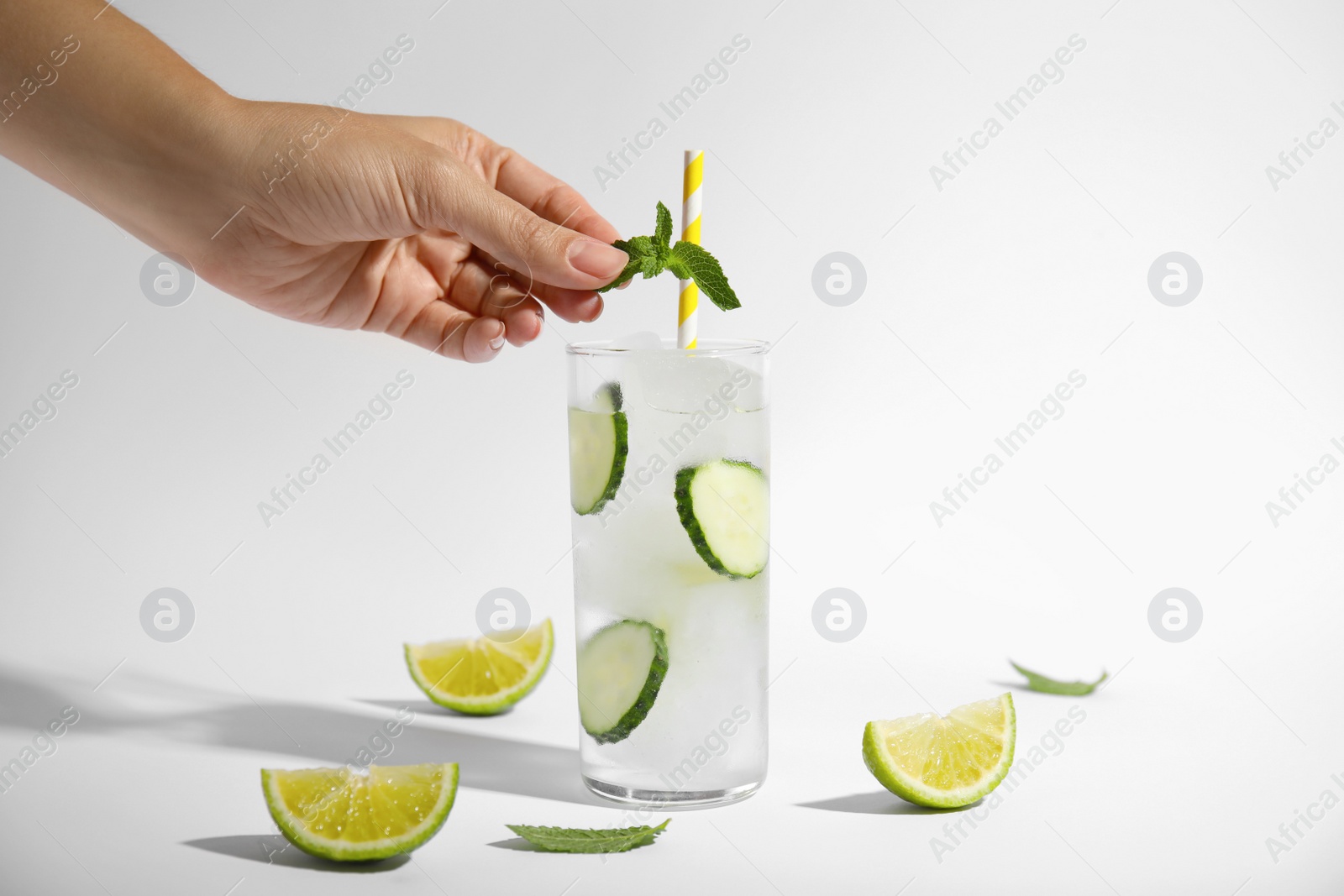 Photo of Woman putting mint into glass of tasty fresh cucumber water and sliced lime on white background, closeup