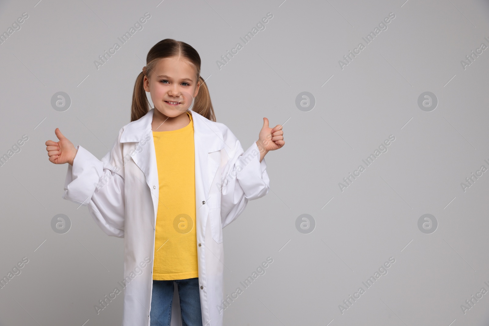 Photo of Little girl in medical uniform showing thumbs up on light grey background. Space for text