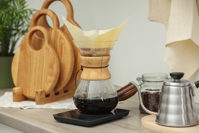 Photo of Making drip coffee. Glass chemex coffeemaker with paper filter, jar of beans and kettle on wooden countertop in kitchen
