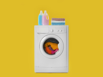 Modern washing machine with laundry, stack of towels and detergents on yellow background