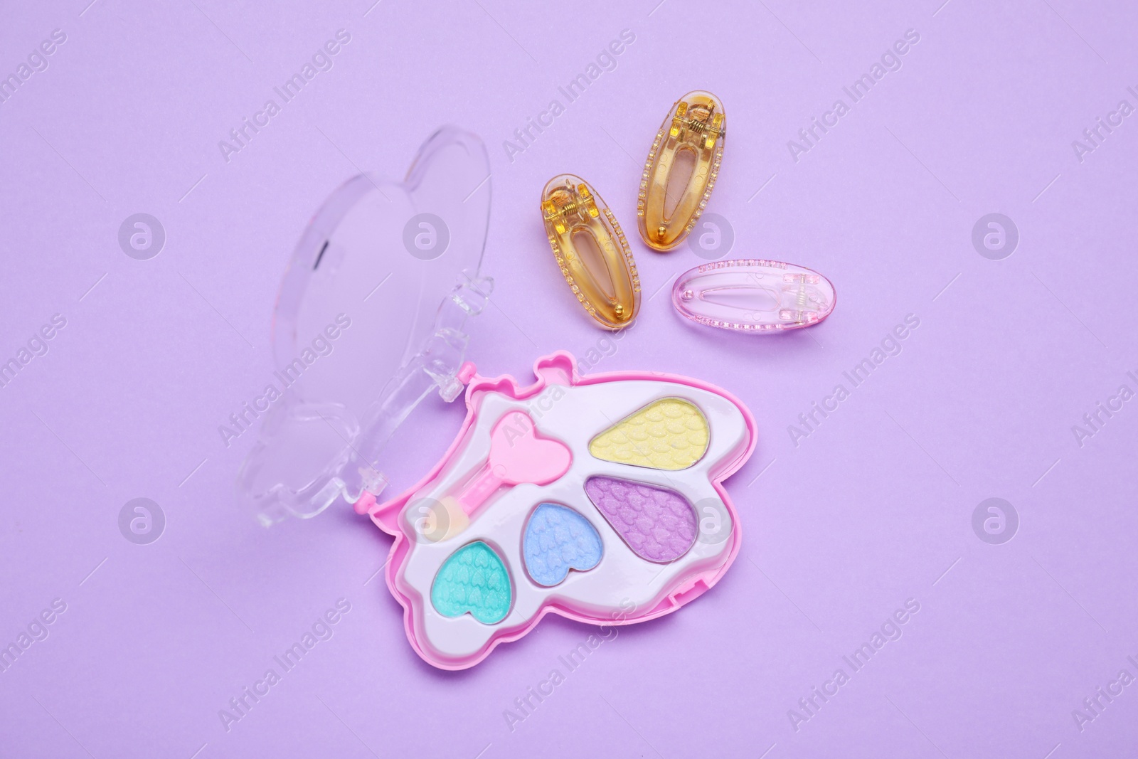 Photo of Children's eye shadow palette with brush and hair clips on violet background, flat lay