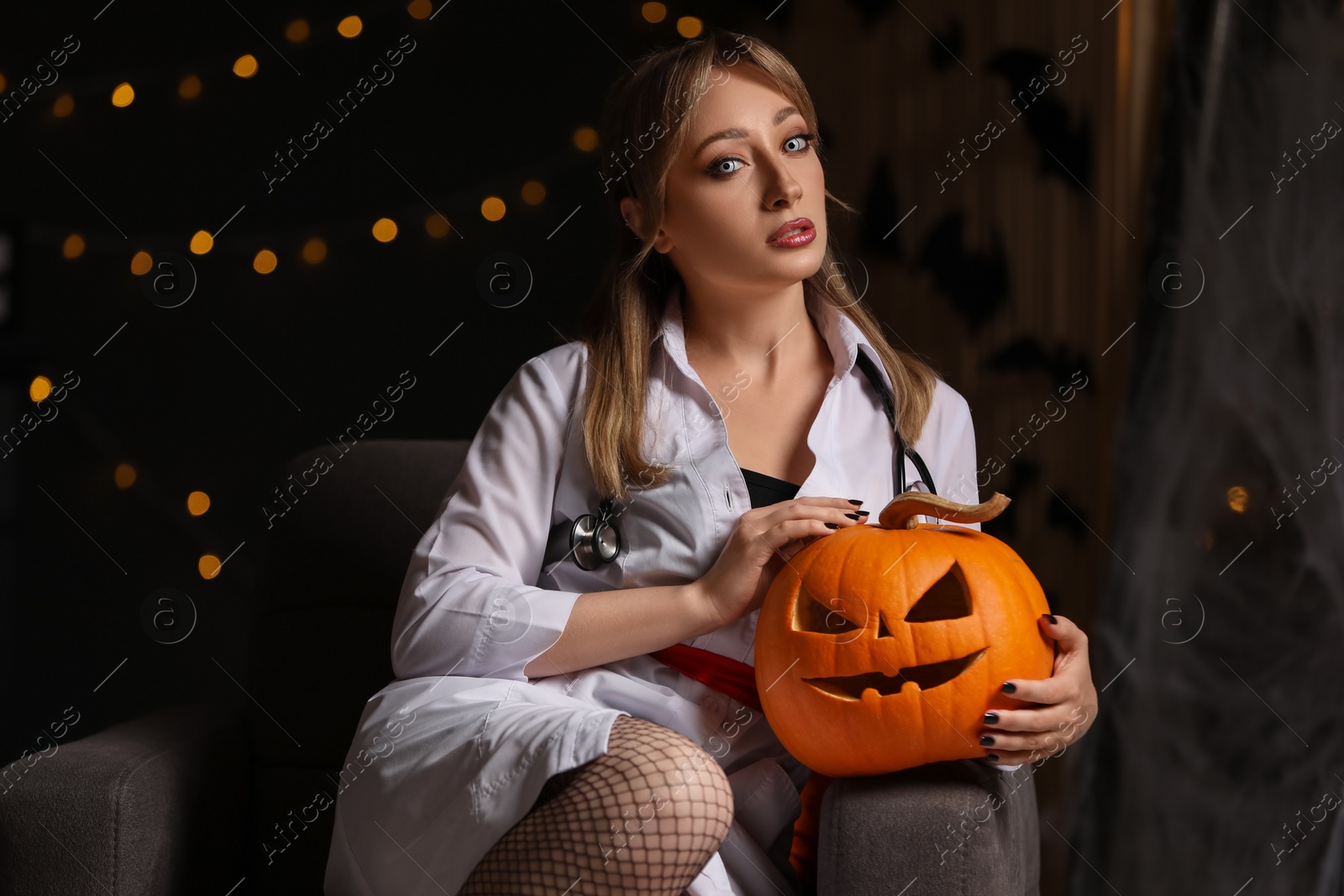 Photo of Woman in scary nurse costume with carved pumpkin against blurred lights indoors. Halloween celebration