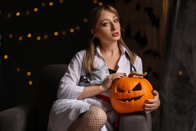 Woman in scary nurse costume with carved pumpkin against blurred lights indoors. Halloween celebration