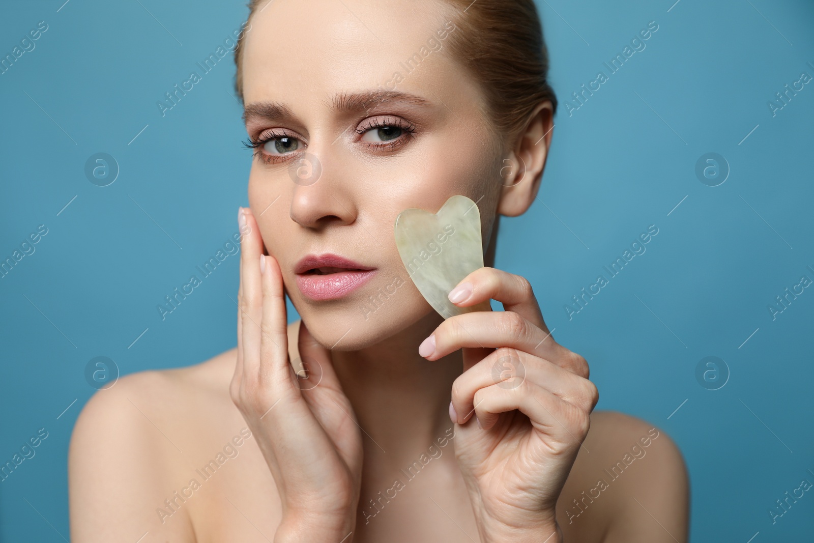 Photo of Beautiful young woman doing facial massage with gua sha tool on blue background, closeup